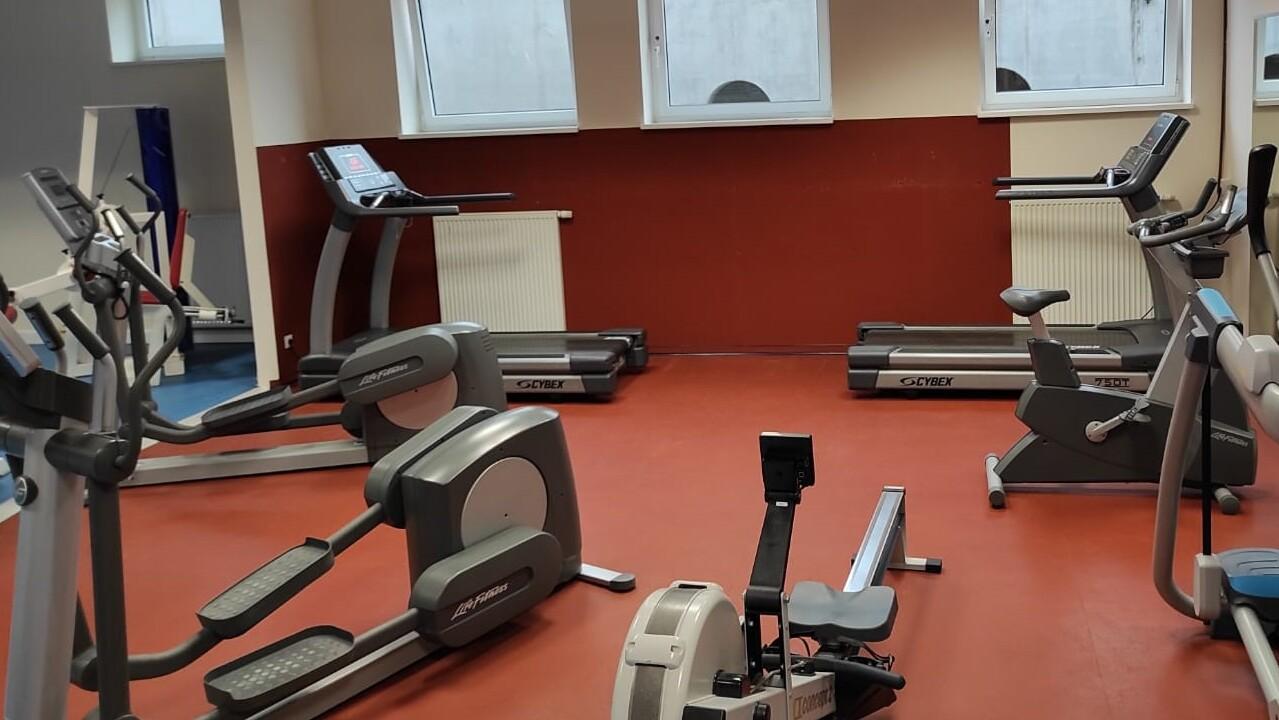 Fitnessstudio mit verschiedenen Trainingsgeräten wie Ergometern, Crosstrainern und einem Rudergerät auf rotem Boden.