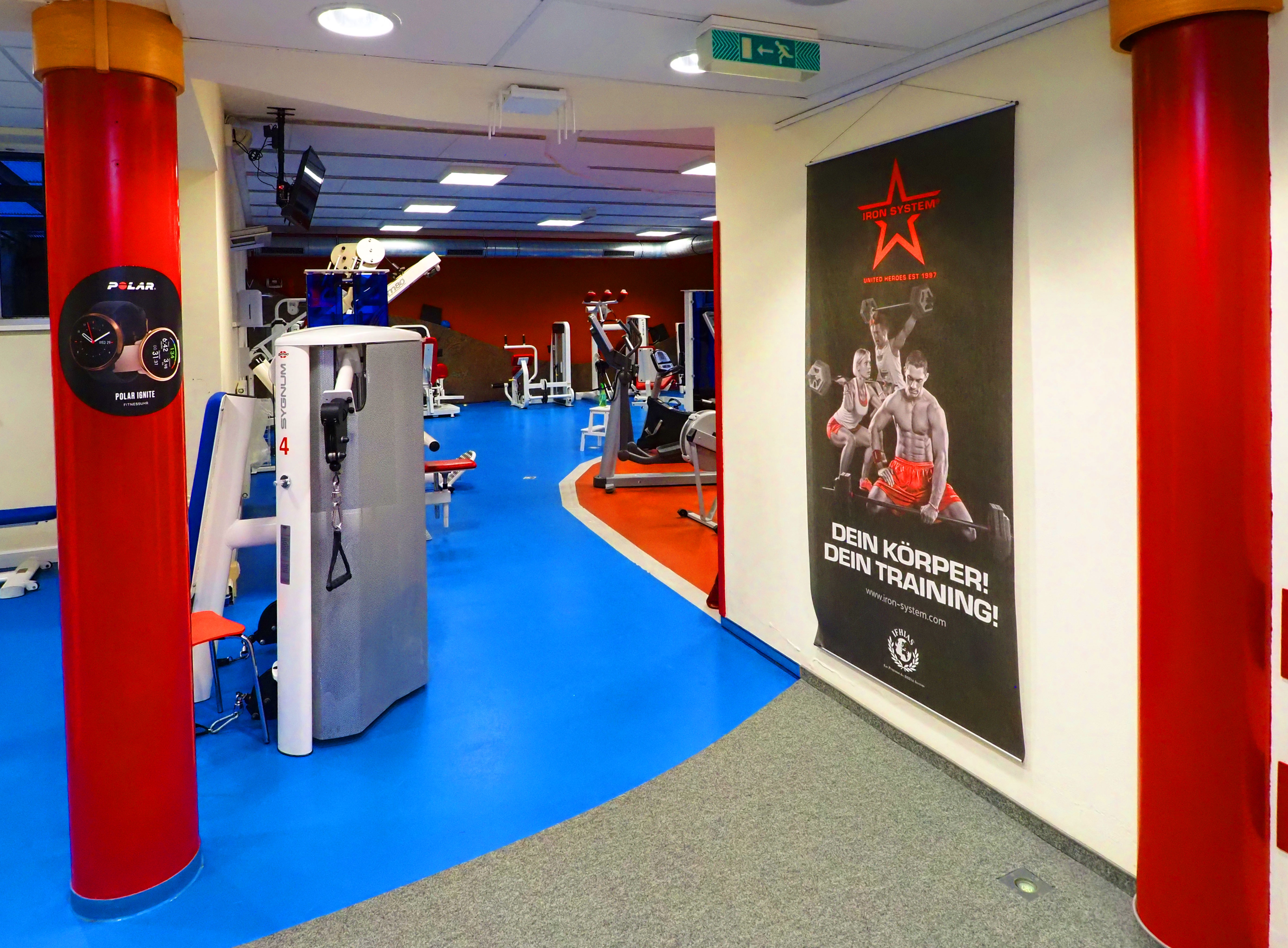 Fitnessstudio mit blauen Böden, Geräten und einem Plakat mit dem Slogan „Dein Körper! Dein Training!“.