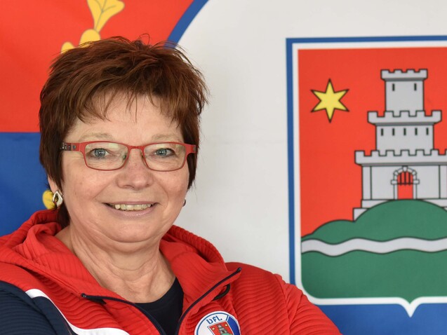 Porträt einer lächelnden Frau in sportlicher Kleidung vor einer Landesflagge mit Wappen im Hintergrund.