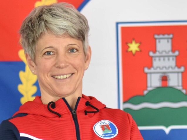 Sportlerin mit kurzen blonden Haaren lächelt vor einer Flagge, die ein Wappen zeigt, und trägt einen sportlichen Hoodie.