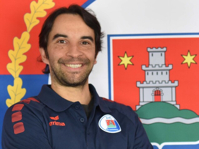 Lächelnder Mann mit dunklen Haaren steht vor einer blauen und roten Fahne mit Wappen, Arme vor der Brust verschränkt.