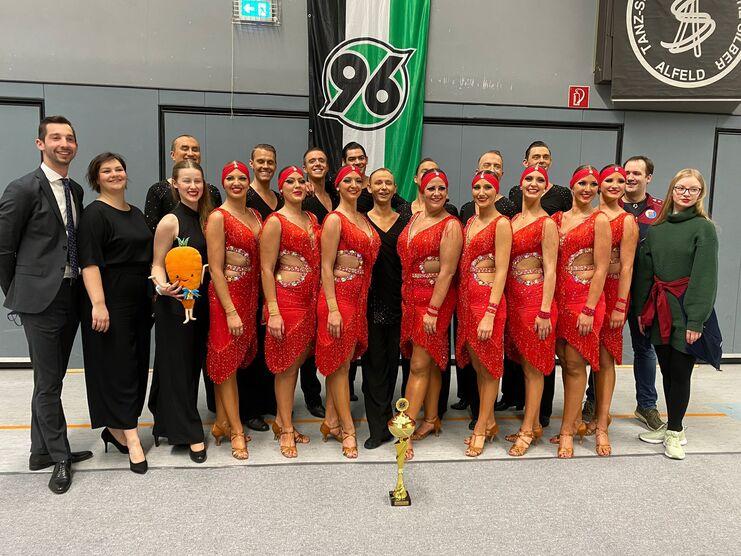 Tanzgruppe in roten Kostümen posiert mit Pokal, umgeben von Zuschauern in einem Sporthallenambiente.