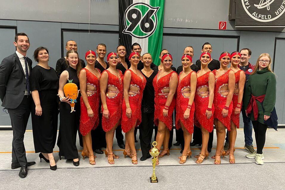 Tanzgruppe in roten Kost&uuml;men posiert mit Pokal, umgeben von Zuschauern in einem Sporthallenambiente.