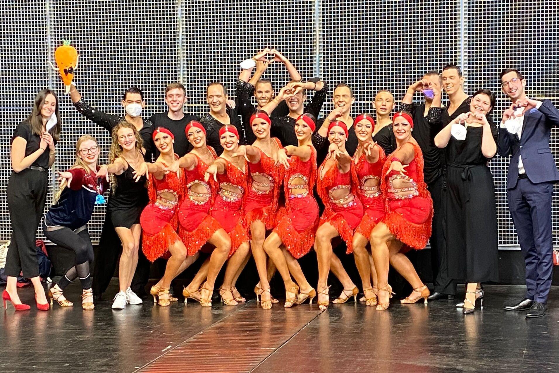 Gruppe von T&auml;nzern in roten Kost&uuml;men posiert auf der B&uuml;hne, umgeben von anderen Teilnehmern in eleganter Kleidung.