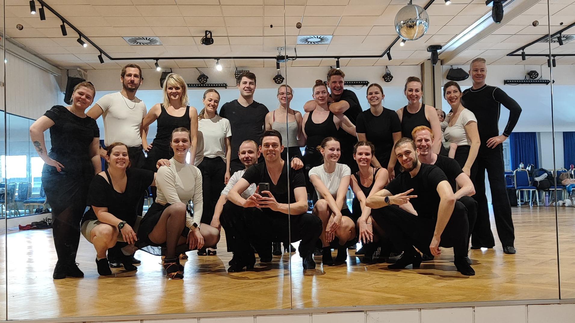 Gruppe von Tänzern posiert in Sportkleidung vor einem großen Spiegel in einem Tanzsaal.