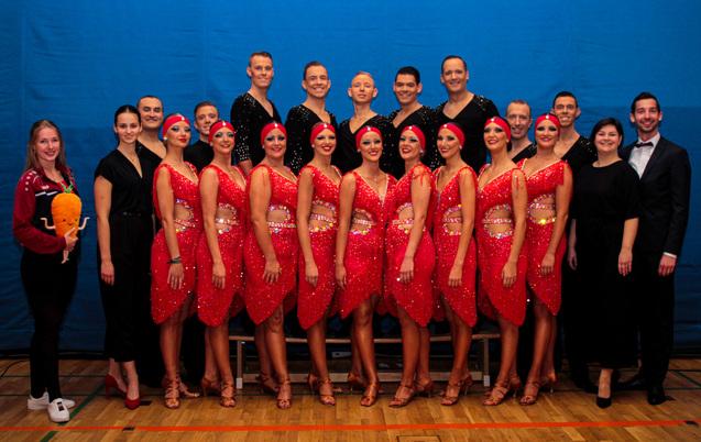 Gruppe von Tänzern in roten Kostümen mit Strass, posierend vor blauem Hintergrund in einer Sporthalle.