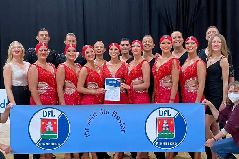 Gruppe von T&auml;nzern in roten Kost&uuml;men posiert mit einem Banner und einem Pokal auf einer Tanzfl&auml;che.