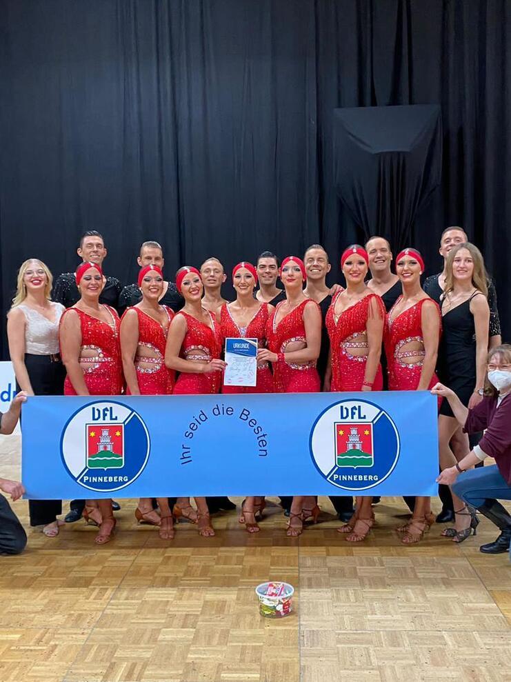 Gruppe von Tänzern in roten Kostümen posiert mit einem Banner und einem Pokal auf einer Tanzfläche.