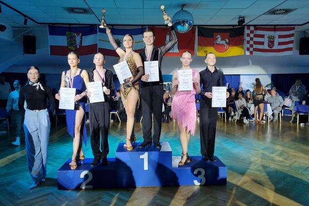 Gruppe von T&auml;nzern auf Podest bei einem Wettbewerb, alle halten Urkunden und Pokale, im Hintergrund sind Zuschauer.