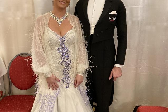 Paar in eleganter Tanzkleidung posiert l&auml;chelnd vor einem wei&szlig;en Vorhang in einem festlichen Raum.