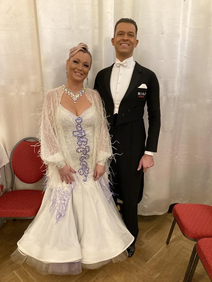 Paar in eleganter Tanzkleidung posiert lächelnd vor einem weißen Vorhang in einem festlichen Raum.
