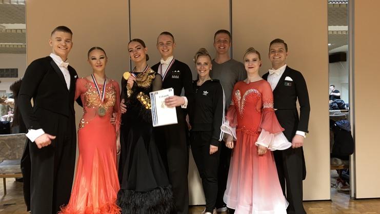 Gruppe von acht Tanzern in eleganter Abendgarderobe, einige mit Medaillen und Urkunden, in einem festlichen Raum.