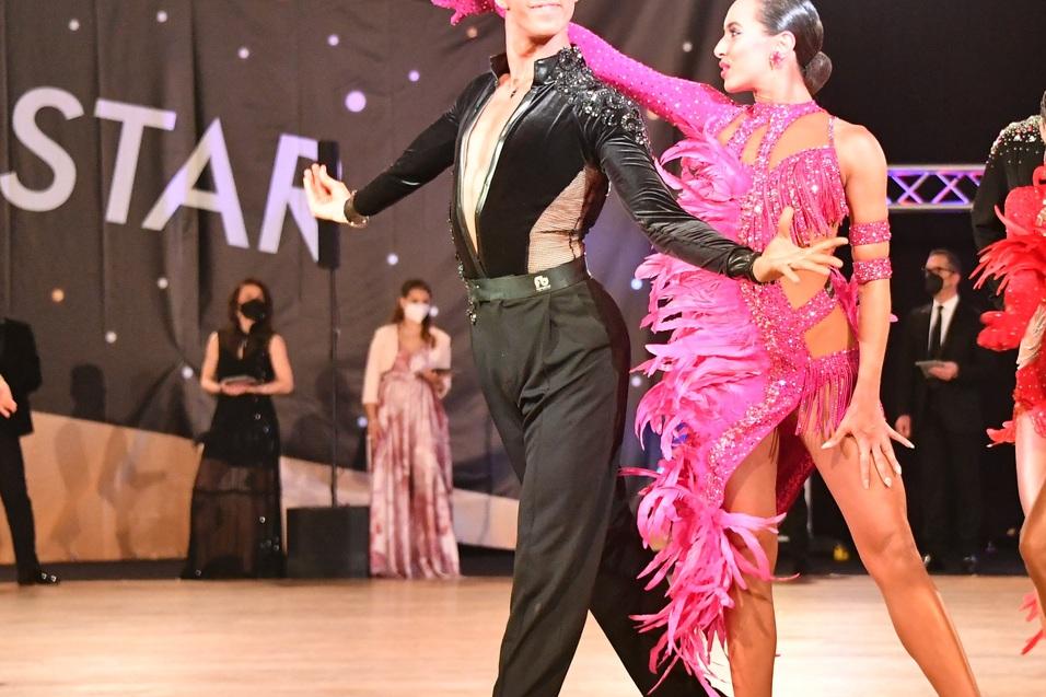 Tanzpaar in eleganter Kleidung in Aktion auf dem Tanzparkett, umgeben von Zuschauern in Abendgarderobe.