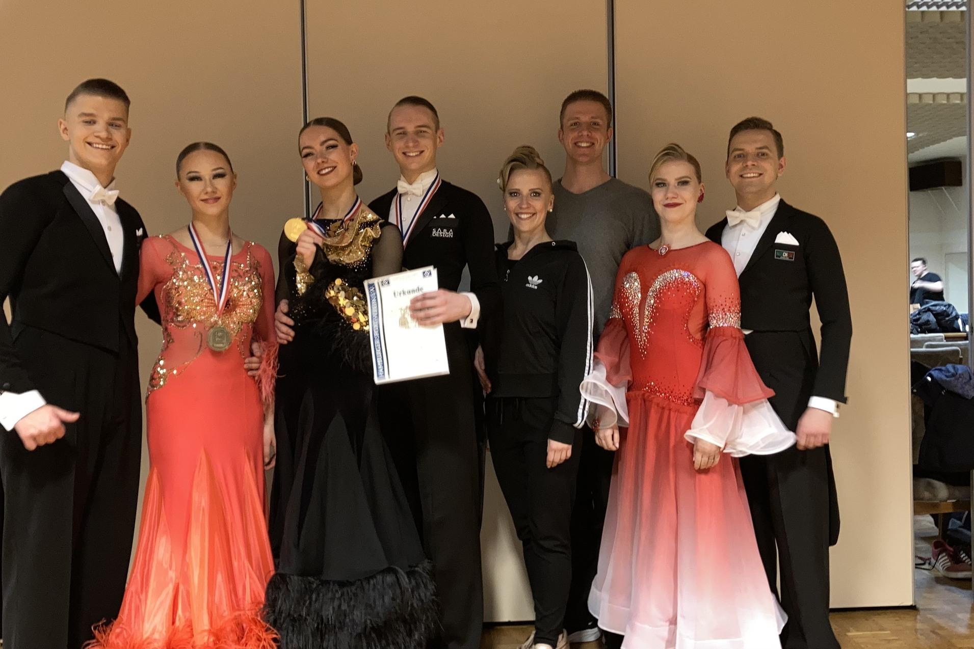Gruppe von acht Tanzern in eleganter Abendgarderobe, einige mit Medaillen und Urkunden, in einem festlichen Raum.