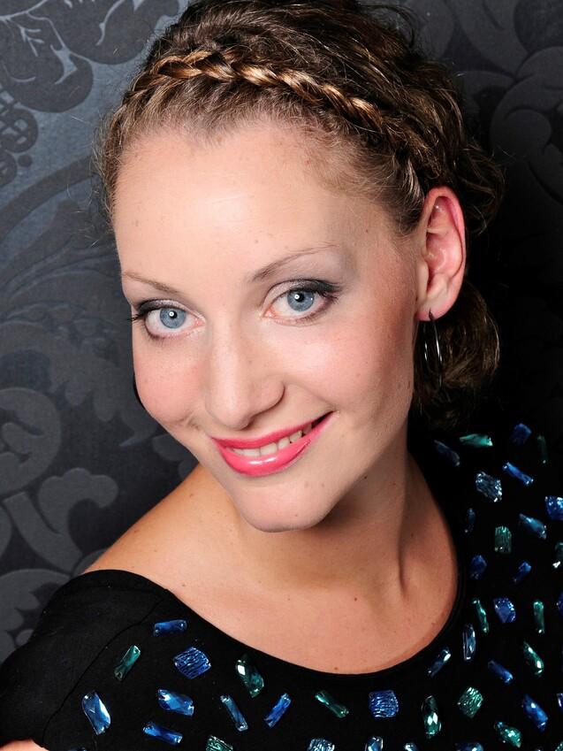 Lächelnde Frau mit blauen Augen und Zopf, trägt ein schwarzes Kleid mit bunten Applikationen vor einem gemusterten Hintergrund.