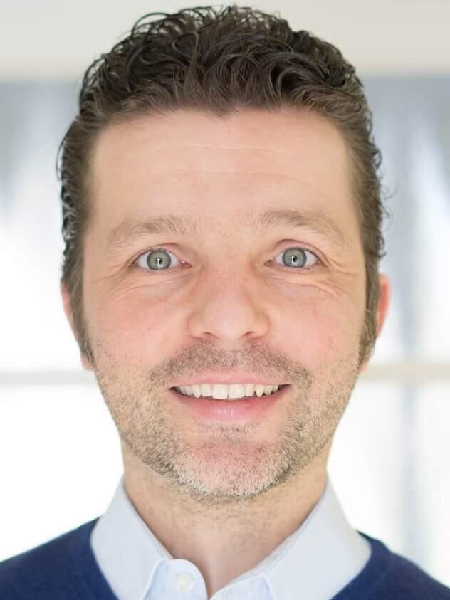 Lächelnder Mann mit kurzem, lockigem Haar, in einem blauen Pullover, vor einem hellen Hintergrund.