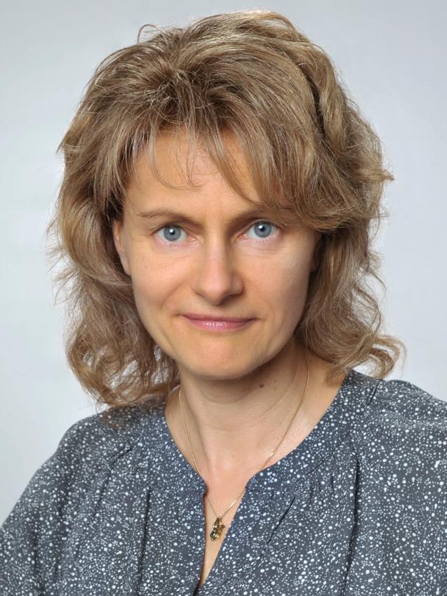 Porträt einer Frau mit lockigem, blonden Haar und blauen Augen, gekleidet in ein graues, gepunktetes Oberteil.