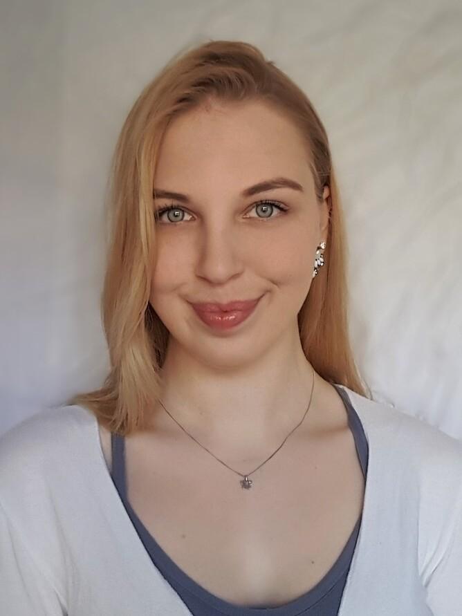 Porträt einer Frau mit blonden Haaren, die ein helles Oberteil trägt, vor einem neutralen Hintergrund.