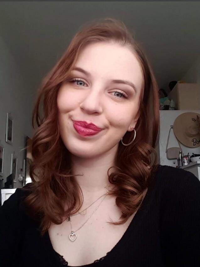Porträt einer lächelnden Frau mit lockigem, braunem Haar, schwarzem Oberteil und Schmuck im Hintergrund.