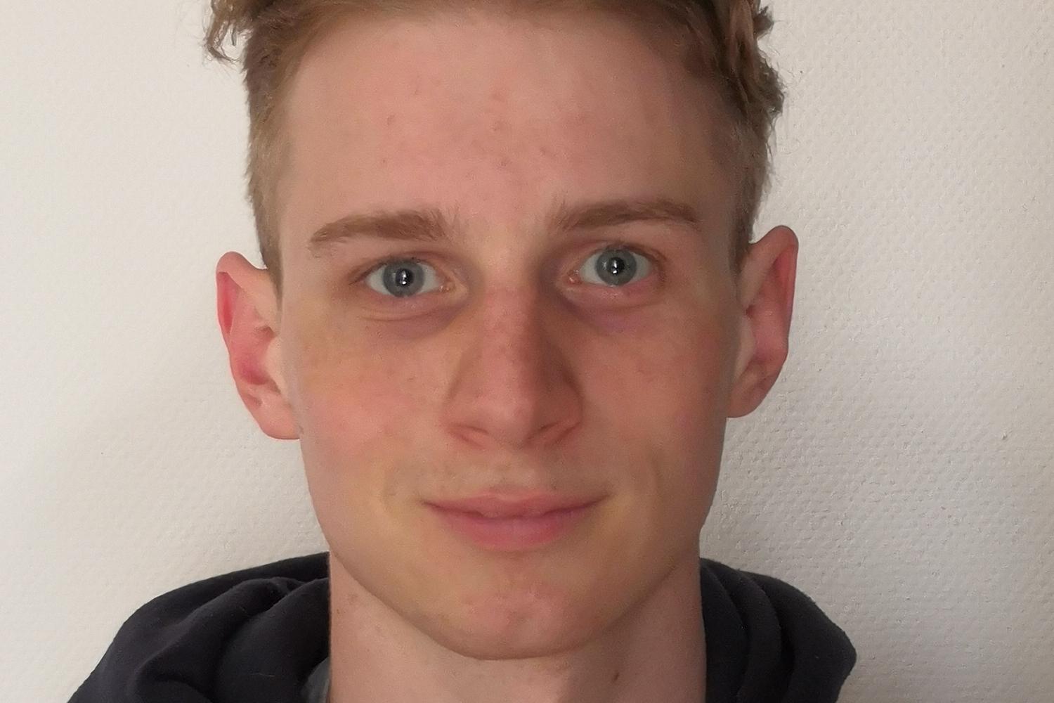 Junger Mann mit kurzem, blondem Haar und blauen Augen, tr&auml;gt einen roten und schwarzen Kapuzenpulli vor einer hellen Wand.