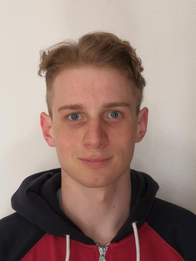 Junger Mann mit kurzem, blondem Haar und blauen Augen, trägt einen roten und schwarzen Kapuzenpulli vor einer hellen Wand.