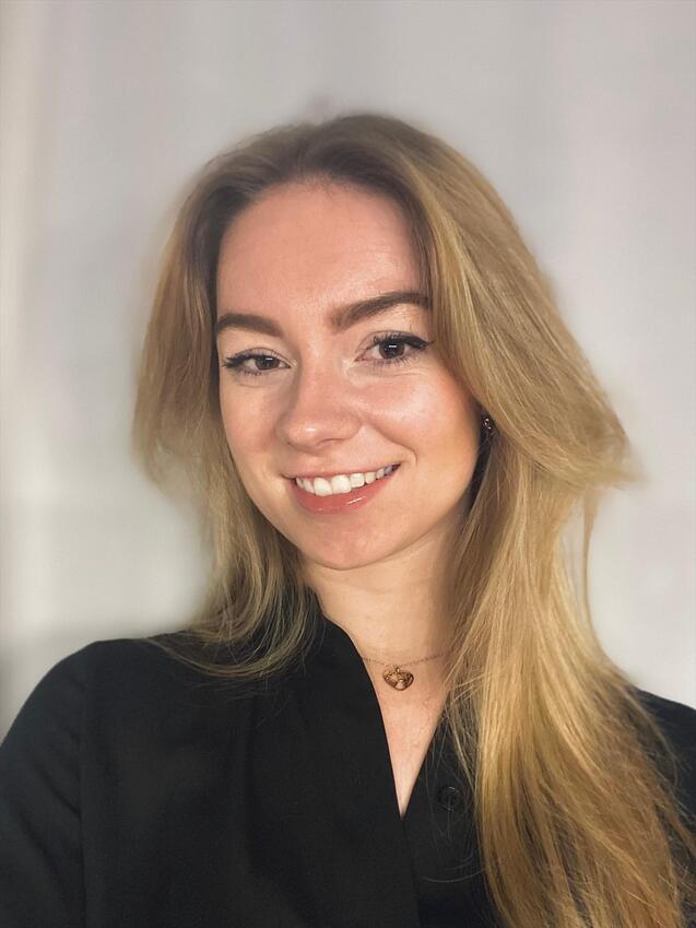 Porträt einer lächelnden Frau mit langen blonden Haaren, schwarzem Oberteil und Herzanhänger vor hellem Hintergrund.