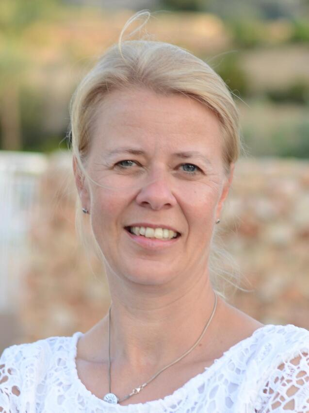 Nahaufnahme einer lächelnden Frau mit blonden Haaren, die ein weißes Oberteil trägt, vor unscharfem Hintergrund.