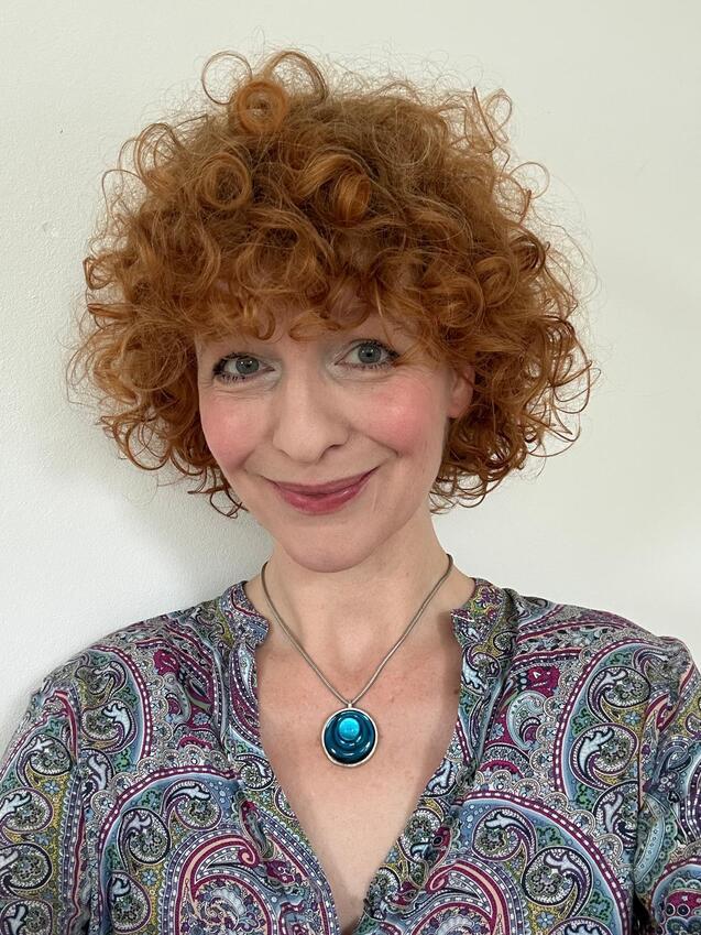 Porträt einer Frau mit lockigem rotem Haar, die ein gemustertes Hemd und eine Halskette mit blauem Anhänger trägt.