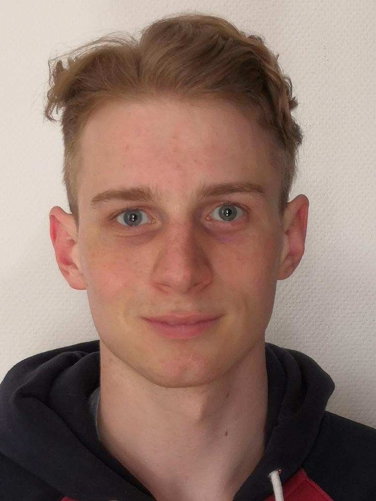 Junger Mann mit kurzem, blondem Haar und blauen Augen, trägt einen roten und schwarzen Kapuzenpulli vor einer hellen Wand.