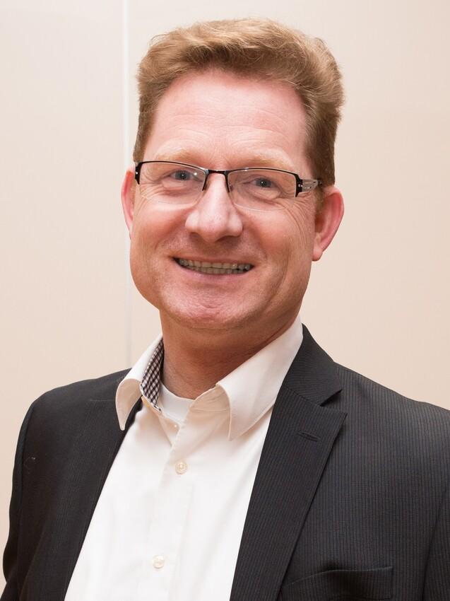 Mann mit Brille und blonden Haaren lächelt, trägt einen schwarzen Anzug und ein weißes Hemd in einem hellen Raum.