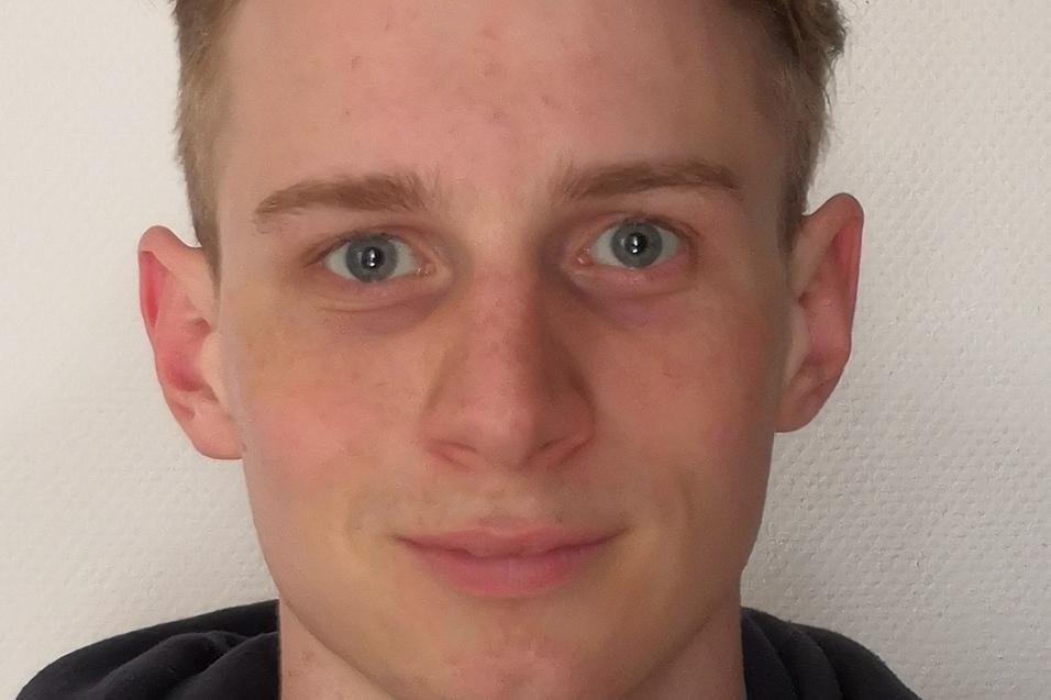 Junger Mann mit kurzem, blondem Haar und blauen Augen, tr&auml;gt einen roten und schwarzen Kapuzenpulli vor einer hellen Wand.