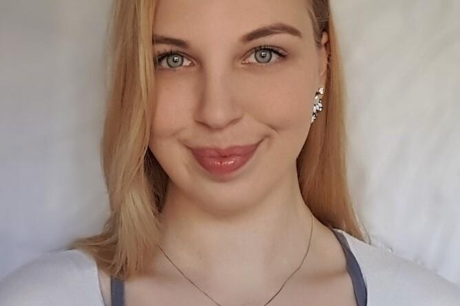 Portr&auml;t einer Frau mit blonden Haaren, die ein helles Oberteil tr&auml;gt, vor einem neutralen Hintergrund.