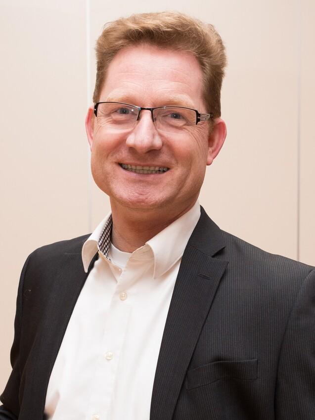 Mann mit Brille und blonden Haaren lächelt, trägt einen schwarzen Anzug und ein weißes Hemd in einem hellen Raum.