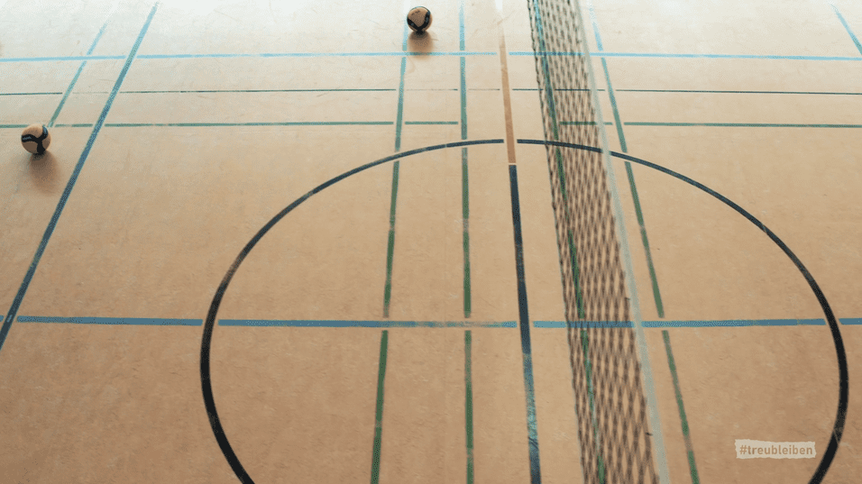Ball liegt auf einem Volleyballfeld mit Linien und Netz. Der Boden ist hellbraun mit bunten Markierungen.