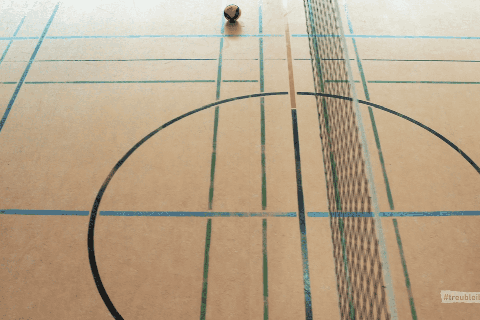 Ball liegt auf einem Volleyballfeld mit Linien und Netz. Der Boden ist hellbraun mit bunten Markierungen.