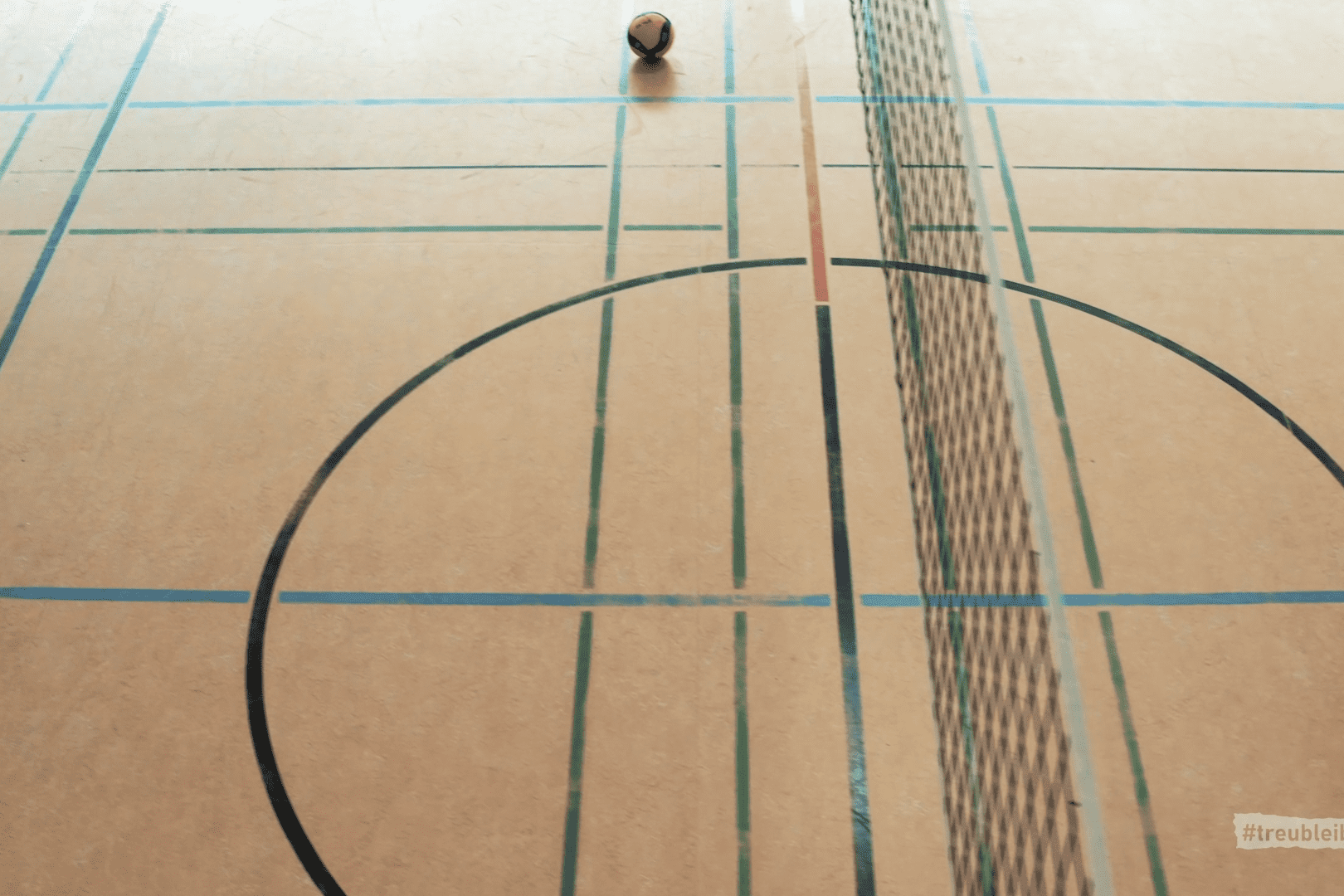 Ball liegt auf einem Volleyballfeld mit Linien und Netz. Der Boden ist hellbraun mit bunten Markierungen.