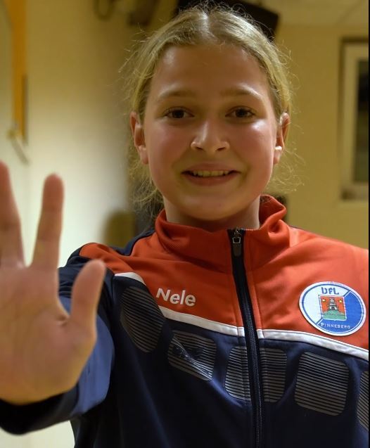 Lächelnde Jugendliche zeigt mit ihrer linken Hand ein Stoppsignal, trägt eine Sportjacke mit Aufschrift „Nele“.