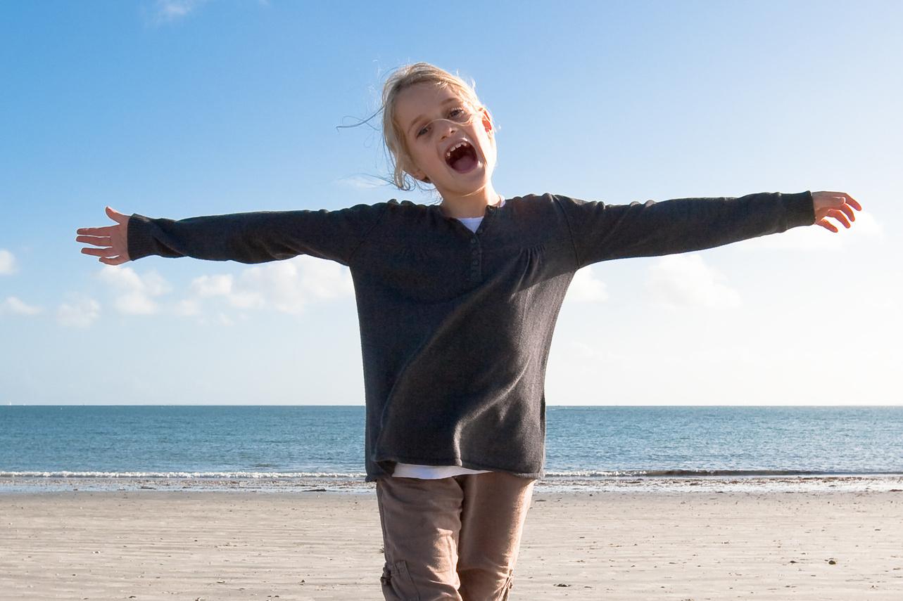 M&auml;dchen steht am Strand mit ausgebreiteten Armen, l&auml;chelt und blickt in die Kamera gegen blauen Himmel und Meer.