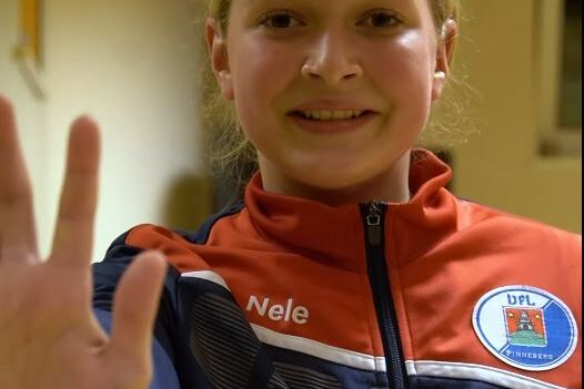 L&auml;chelnde Jugendliche zeigt mit ihrer linken Hand ein Stoppsignal, tr&auml;gt eine Sportjacke mit Aufschrift &bdquo;Nele&ldquo;.