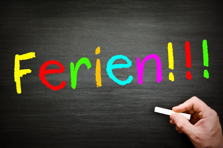 Bunte Schrift &bdquo;Ferien!!!&ldquo; auf einer Tafel, mit einer Hand, die mit Kreide schreibt.