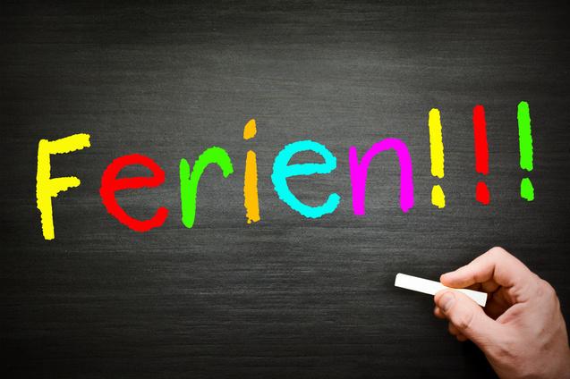 Bunte Schrift &bdquo;Ferien!!!&ldquo; auf einer Tafel, mit einer Hand, die mit Kreide schreibt.