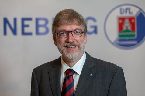 Portrait eines Mannes mit Brille, grauem Anzug und gestreifter Krawatte, lächelnd vor einem neutralen Hintergrund.