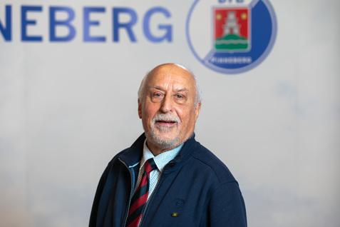 Porträt eines älteren Mannes mit grauen Haaren, der einen blauen Pullover und gestreifte Krawatte trägt, vor neutralem Hintergrund.
