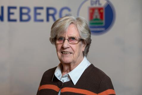 Ältere Frau mit Brille lächelt vor einem grauen Hintergrund mit dem Logo eines Vereins. Sie trägt einen braunen Pullover.