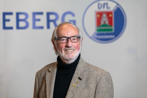 Porträt eines älteren Mannes mit Brille und grauen Haaren, in einem braunen Blazer, vor einem neutralen Hintergrund.