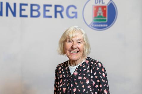 Lächelnde ältere Frau mit blonden Haaren und blumenmusterbluse vor einem verschwommenen Hintergrund mit Schriftzug.