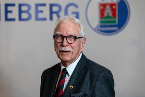 Älterer Mann mit Brille und weißen Haaren, lächelnd vor einem Hintergrund mit dem Schriftzug „Neuenberg“.