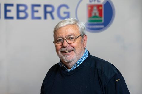 Lächelnder älterer Mann mit Brille, in blauer Pullover, vor einer hellen Hintergrundwand mit Schriftzug.