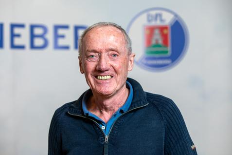 Porträt eines lächelnden älteren Mannes in blauem Pullover mit Reißverschluss, vor einem Hintergrund mit Logos.