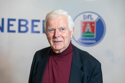 Porträt eines älteren Mannes mit grauen Haaren, schwarzer Jacke und rotem Rollkragenpullover vor einem DFL-Hintergrund.